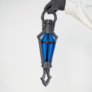 May include: A grey and blue lantern with a cross design. The lantern has a blue centre with a brown cross and is held by a gloved hand. The lantern has a handle and a decorative bottom.