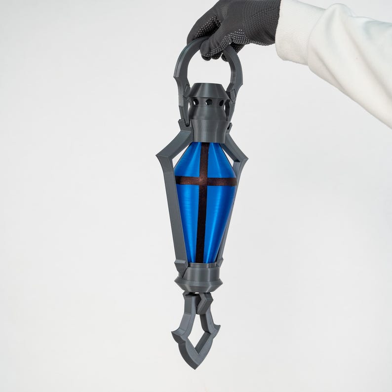 May include: A grey and blue lantern with a cross design. The lantern has a blue centre with a brown cross and is held by a gloved hand. The lantern has a handle and a decorative bottom.