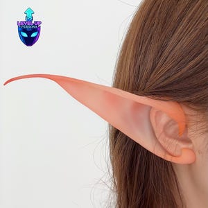 May include: A pair of translucent, peach-colored elf ear prosthetics are attached to a person's ear. The prosthetic extends upwards and outwards, creating a pointed, elven appearance. The Level Up Persona logo is visible in the upper left corner.