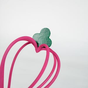 K&ouml;nnte beinhalten: Nahaufnahme eines herzf&ouml;rmigen Objekts in Pink mit einem gr&uuml;nen Kleeblatt-Akzent. Das Objekt hat ein glattes, geschwungenes Design und befindet sich vor einem schlichten wei&szlig;en Hintergrund. Die Herzform wird durch drei gebogene Linien gebildet.