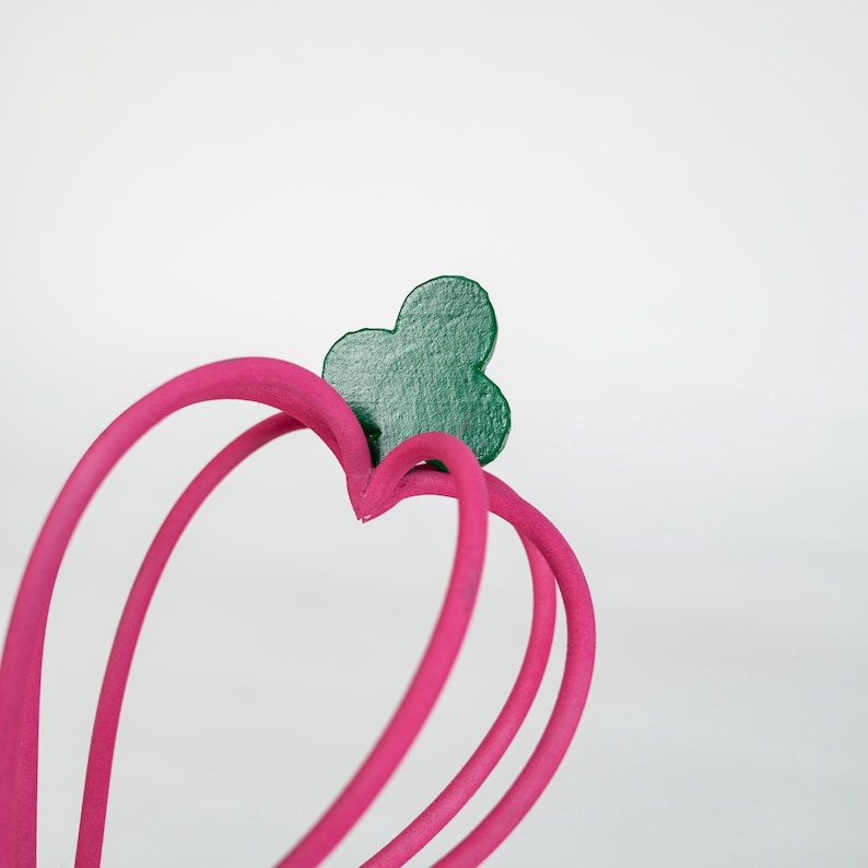 K&ouml;nnte beinhalten: Nahaufnahme eines herzf&ouml;rmigen Objekts in Pink mit einem gr&uuml;nen Kleeblatt-Akzent. Das Objekt hat ein glattes, geschwungenes Design und befindet sich vor einem schlichten wei&szlig;en Hintergrund. Die Herzform wird durch drei gebogene Linien gebildet.