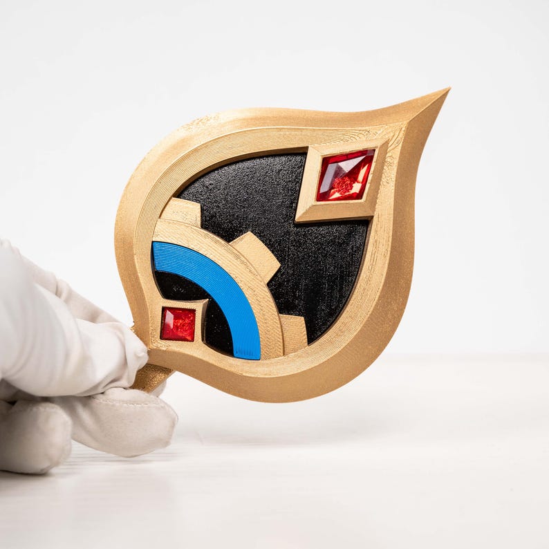 May include: A gold-coloured, leaf-shaped decorative item with a black centre. It features a gear design with blue accents and two red square gems. The item is held by a gloved hand against a white background.