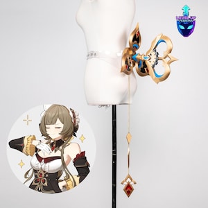 May include: A gold and blue cosplay accessory, possibly a key or emblem, attached to a clear belt. The design features intricate details and a dangling chain with diamond and gem-shaped pendants. A cartoon image is in the lower left corner.