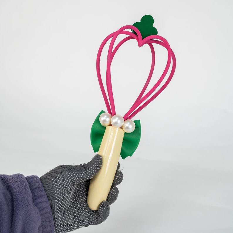 K&ouml;nnte beinhalten: Ein skurriler Zauberstab mit einem herzf&ouml;rmigen rosa Oberteil und einem gr&uuml;nen Kleeblatt. Der Zauberstab hat einen cremefarbenen Griff, eine gr&uuml;ne Schleife und drei wei&szlig;e perlenartige Perlen. Der Zauberstab wird von einer behandschuhten Hand gehalten.