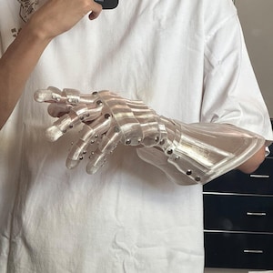 May include: A clear, articulated robotic hand with silver rivets. The hand is designed with segmented fingers and a wrist guard, suggesting a futuristic or mechanical theme. The material appears to be translucent plastic.