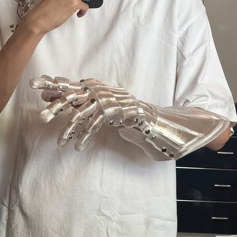 May include: A clear, articulated robotic hand with silver rivets. The hand is designed with segmented fingers and a wrist guard, suggesting a futuristic or mechanical theme. The material appears to be translucent plastic.