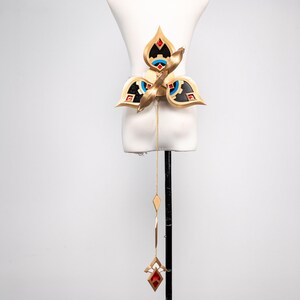 May include: A gold-coloured belt with a floral design, featuring red, black, and blue accents. A gold chain with diamond-shaped and geometric pendants hangs from the belt, culminating in a red jewel pendant. The belt is displayed on a white mannequin.