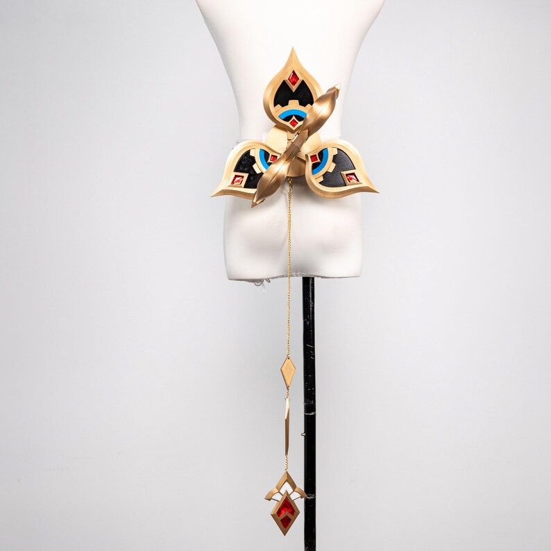 May include: A gold-coloured belt with a floral design, featuring red, black, and blue accents. A gold chain with diamond-shaped and geometric pendants hangs from the belt, culminating in a red jewel pendant. The belt is displayed on a white mannequin.