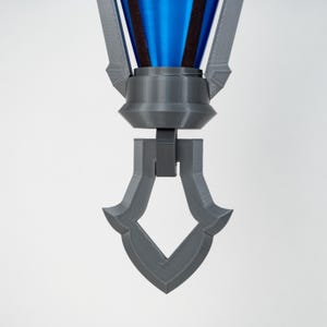 May include: A 3D-printed, grey and blue decorative object. The top section is blue with a dark brown band, connected to a grey, geometric base. The base has a diamond-shaped cutout at the bottom. The object is set against a white background.