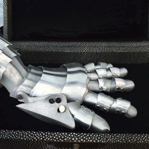 May include: A silver-colored articulated hand, possibly a costume piece or sculpture, with segmented fingers and a detailed design. The hand is resting on a black, textured surface, suggesting a display or storage setting.