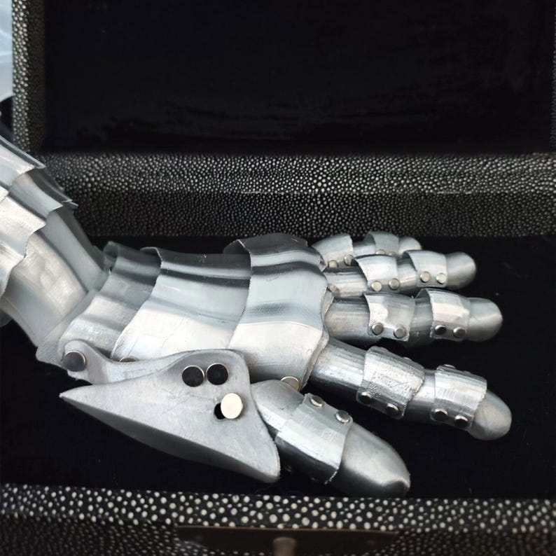 May include: A silver-colored articulated hand, possibly a costume piece or sculpture, with segmented fingers and a detailed design. The hand is resting on a black, textured surface, suggesting a display or storage setting.