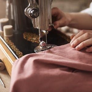 May include: A vintage black sewing machine with gold accents is in use. A person's hands guide a piece of pink fabric under the needle. Spools of thread and sewing tools are visible on the wooden surface.