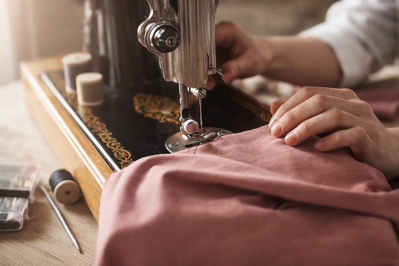 Peut inclure: Une machine &agrave; coudre noire vintage avec des d&eacute;tails dor&eacute;s est en cours d'utilisation. Les mains d'une personne guident un tissu rose sous l'aiguille. Des bobines de fil et des outils de couture sont visibles.