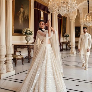 May include: A woman in a white, embellished lehenga with a sheer cape poses in a grand hall. The skirt is detailed with reflective accents. A man in a cream-colored kurta walks in the background. Chandeliers and artwork adorn the space.