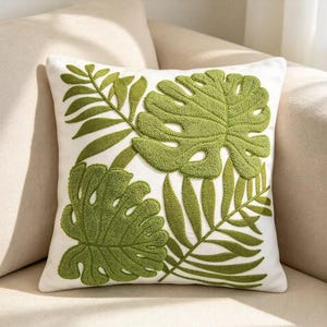 May include: A square throw pillow featuring a white base with raised, textured green embroidery of monstera and palm leaves. The design covers the front surface with detailed, tufted botanical patterns in varying shades of green.