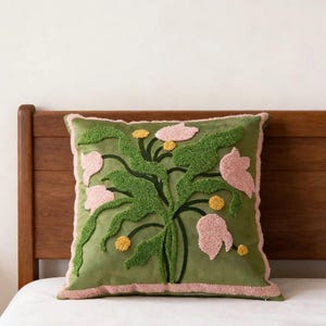 Pode incluir: Uma almofada decorativa quadrada verde-oliva com um design floral. O design apresenta folhas verdes, flores rosa e detalhes amarelos. A almofada tem uma borda rosa e está colocada sobre uma superfície branca.