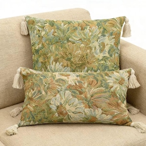 May include: Two rectangular throw pillows featuring a woven floral tapestry pattern in shades of sage green, olive, and muted gold. Each corner of the pillows is finished with a small, cream-colored fabric tassel for added texture.