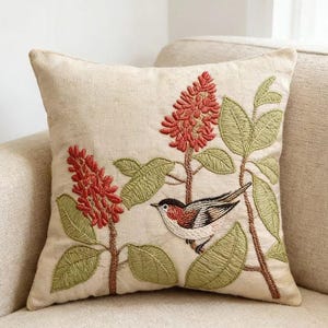 Può includere: Cuscino quadrato beige con design ricamato. Il design presenta un uccello appollaiato su un ramo con fiori rossi e foglie verdi. Il cuscino è su un divano beige.