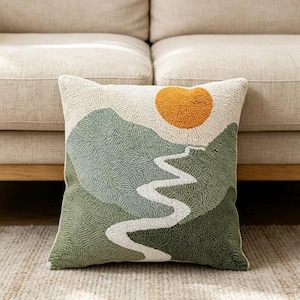 May include: A square throw pillow featuring a textured punch needle design of a landscape. The scene depicts rolling hills in shades of sage and forest green, a winding white path leading upward, and a solid mustard yellow sun against a cream background.