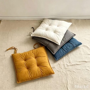 Puede incluir: Una pila de cuatro cojines de asiento cuadrados con detalles capitoné y lazos de tela en las esquinas. Los cojines son de color amarillo mostaza, crema, gris carbón y azul marino, cada uno con un acabado suave y texturizado.