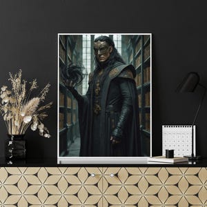 Op de afbeelding: Een ingelijste print met een persoon in een zwarte mantel en een gouden masker, die een donkere bol vasthoudt. De achtergrond toont een bibliotheek met boekenplanken. De print wordt weergegeven op een zwart oppervlak.