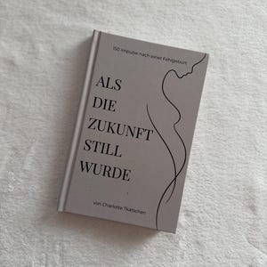 May include: A grey hardcover book with the title "ALS DIE ZUKUNFT STILL WURDE" in large, dark letters. The cover also features a line drawing of a woman's profile and torso. The author's name, Charlotte Tkatsch, is printed at the bottom of the book.