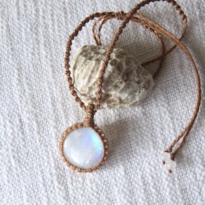 May include: A round, iridescent moonstone cabochon set in a brown macrame cord pendant. The pendant hangs from a matching brown cord necklace that features small, clear beads at the ends of the adjustable sliding closure mechanism.