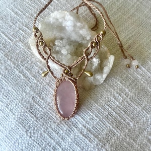 May include: A macrame necklace featuring a central oval rose quartz cabochon, woven with light tan cord. The design includes small metallic gold-toned beads and two teardrop-shaped charms, finished with adjustable braided cords and small end beads.