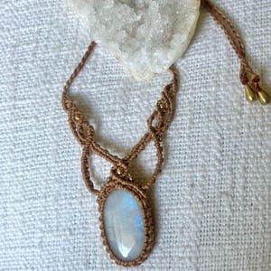 May include: A macrame necklace featuring a central oval moonstone cabochon with a blue iridescent sheen. The pendant is encased in light brown cord with intricate knotting and small gold-toned beads. The necklace includes adjustable cord ends.