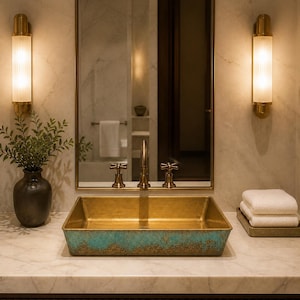 May include: A rectangular vessel sink with a metallic gold interior and a textured, oxidized turquoise exterior finish. The sink is positioned on a white marble countertop with a brass faucet, a dark vase with greenery, and a stack of white towels.