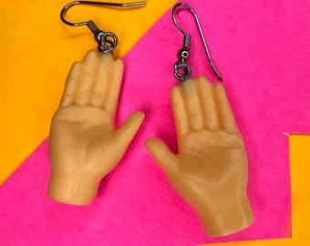 May include: A pair of novelty earrings featuring miniature hand sculptures in a flesh-toned color. Each earring has a silver-colored hook and a coiled wire detail connecting the hand to the hook. The background is a vibrant mix of pink and yellow.