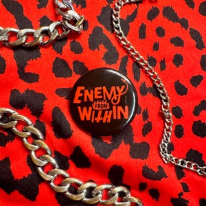 May include: A black button with the words "ENEMY FROM WITHIN" in red lettering and a small red devil tail. The button is on a red fabric with black leopard spots, with silver chains.
