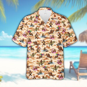Può includere: Camicia a maniche corte con un motivo ripetuto di Bigfoot, dinosauri e cavalli su una spiaggia. La camicia ha uno sfondo beige con palme e altri elementi della spiaggia. I Bigfoot sono impegnati in varie attività sulla spiaggia.