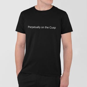 May include: Black t-shirt with the white text "Perpetually on the Cusp" across the chest. The shirt is a crew neck style and appears to be made of a soft material. The person wearing the shirt has their hands in their pockets.