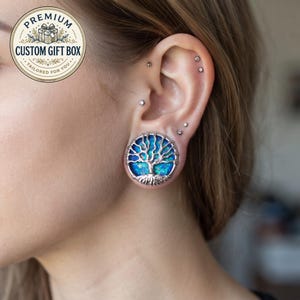 Peut inclure: Un écarteur d'oreille circulaire arborant un motif d'arbre de vie argenté sur un fond bleu irisé vibrant. Le bijou est porté dans le lobe de l'oreille, accompagné de plusieurs petites boucles d'oreilles à tige argentées sur le cartilage.