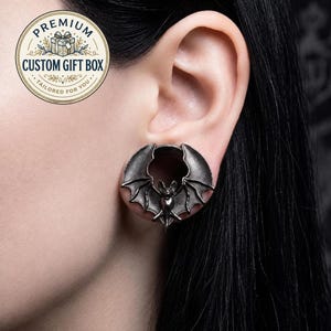 May include: A dark metallic ear plug featuring a detailed bat design with outstretched wings. The jewelry has a circular center opening and a textured, antiqued finish. The bat is positioned centrally, with its wings extending to the edges of the plug.