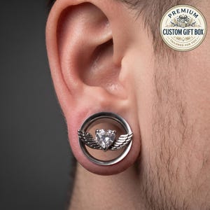 Könnte beinhalten: Ein silberfarbener, kreisförmiger Ohr-Tunnel mit einem zentralen, herzförmigen, klaren Edelstein, der von zwei detaillierten Metallflügeln flankiert wird. Der Schmuck ist in einen glatten, polierten Metallring eingefasst.