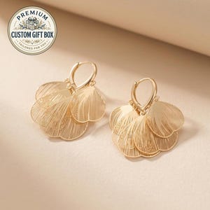 May include: A pair of gold-toned hoop earrings featuring multiple dangling, fan-shaped charms with a delicate, fine-lined texture. The circular hoops have a secure clasp mechanism. A circular logo in the corner reads PREMIUM CUSTOM GIFT BOX.