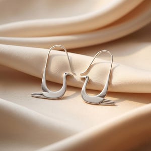 May include: A pair of silver-colored swan earrings. Each earring features a swan design with a curved body, a long neck, and a small beak. The earrings are set against a soft, neutral-toned background.