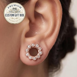 Puede incluir: Un dilatador de oreja en oro rosa con un diseño circular y borde festoneado. El borde exterior está engastado con una serie de pequeñas piedras redondas y transparentes. El centro está hueco. Un logo circular arriba a la izquierda dice PREMIUM CUSTOM GIFT BOX TAILORED FOR YOU.
