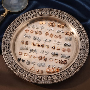 May include: A collection of small metal stud earrings in various shapes including stars, hearts, flowers, triangles, and circles, finished in gold, silver, and rose gold tones, arranged on a round, ornate, metallic decorative tray with a raised rim.