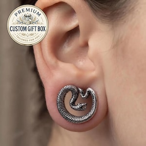 Puede incluir: Un expansor de oreja plateado con un diseño de serpiente enroscada con escamas detalladas y la boca abierta. La joya circular está insertada en el lóbulo. Una insignia circular en la esquina superior izquierda muestra el texto PREMIUM CUSTOM GIFT BOX.