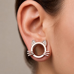 May include: A rose gold-toned ear plug featuring a cat-shaped design with a circular center opening. The cat ears are accented with small, clear, sparkling stones, and the piece includes three horizontal whiskers on each side of the circular frame.