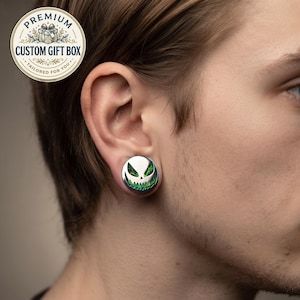 Può includere: Un dilatatore per orecchie circolare con una montatura in metallo color argento e un design stilizzato a faccia sorridente, con occhi triangolari appuntiti e una bocca frastagliata riempita da un inserto verde iridescente e scintillante.