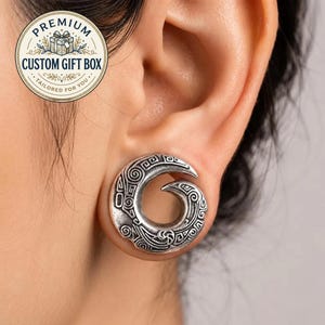 May include: A silver-toned ear gauge featuring a spiral design with intricate engraved patterns. The circular jewelry has a hollow center and a textured, oxidized finish that highlights the detailed carvings across the entire surface of the piece.