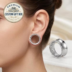 Puede incluir: Un expansor de oreja plateado de forma circular, con un borde exterior liso y una superficie interior texturizada y martillada. El expansor se muestra tanto insertado en el lóbulo de la oreja como por separado, destacando el acabado metálico.