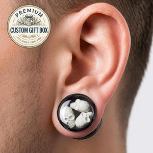 Op de afbeelding: Een ronde oorplug met een zwarte rand en een doorzichtige voorkant met drie kleine, witte, driedimensionale schedels erin. De plug zit in een oorlel. Een rond embleem in de hoek bevat de tekst PREMIUM CUSTOM GIFT BOX.