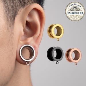 Peut inclure: Un tunnel d'oreille argenté avec un petit anneau circulaire à la base, porté au lobe. Trois autres tunnels d'oreille sont présentés séparément en tons doré, noir et or rose, chacun doté d'un petit anneau circulaire assorti à la base.