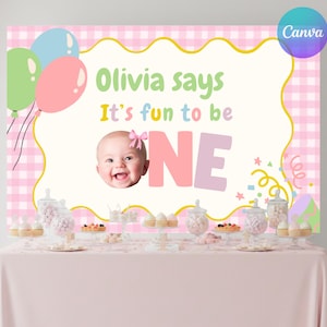 May include: A pink and white gingham backdrop with the words "Olivia says It's fun to be ONE" and a photo of a baby girl. Balloons and a table with treats complete the scene, perfect for a first birthday celebration.