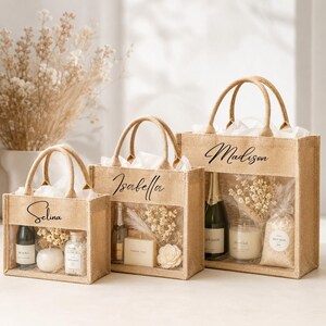 May include: Three jute tote bags with clear front panels, each personalized with a name in elegant script: Selina, Isabella, and Madison. The bags contain gifts like candles, bath salts, and champagne, with dried floral accents.